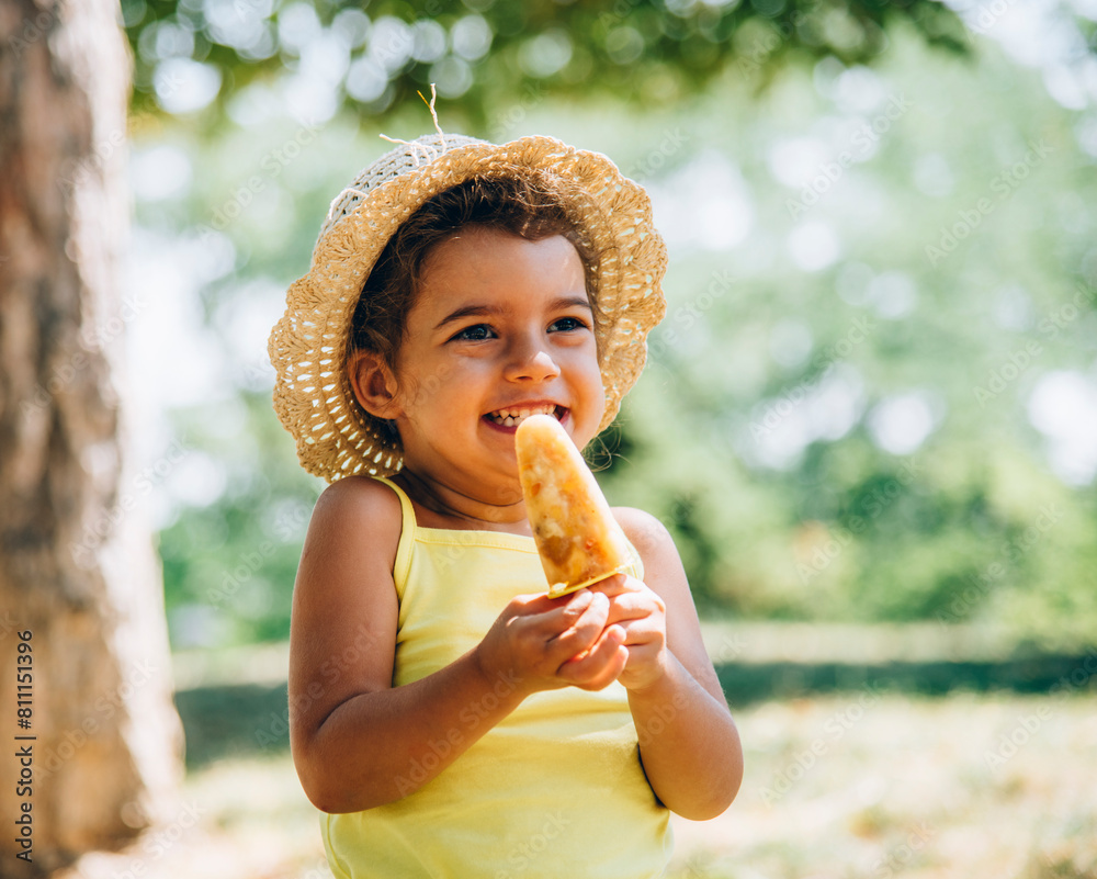 Cute happy little girl in yellow dress and hat eating fruit ice cream. Summer fun child playing in park. Summertime vibrant color background. Cute happy little girl in yellow dress and hat eating fruit ice cream. Summer fun child playing in park. Summertime vibrant color background.
