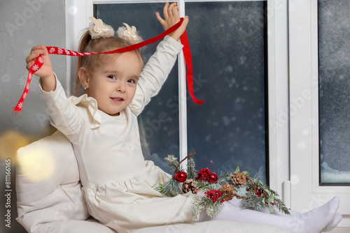 Little happy girl sitting on the windowsill playing with Christmas decor.