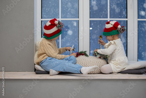 Little children brother and sister in Christmas hats play while sitting on a window with Christmas decor.