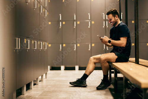 Side view of a smiling fitness man, sitting on the bench in the locker room, using a mobile phone, just finished his training. (ID: 811193176)