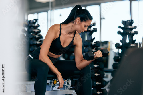 The gym girl working on her biceps, holding a dumbbell, and sitting on the bench, at the gym.