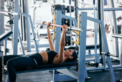 A strong girl doing exercise for her chest, holding a barbell, laying on the bench, at the gym.