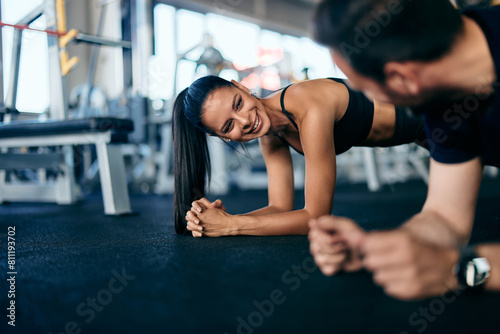 Gym couple doing abs together, holding a plank, looking at each other, smiling.