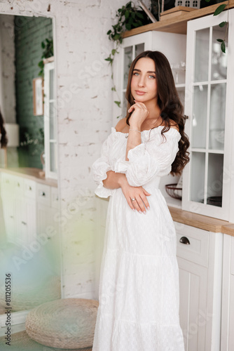 Women's day. Romantic attractive young woman 20-25 years with long curly hair in white dress is poses with crossed hands and looks at camera at modern stylish interior of home kitchen on background.