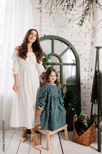 Mother's day. Elegant young mom and little cute daughter sit and smile, embrace on wooden chair at living room at home, look at camera. Trust, support and love between mommy and child. Children day