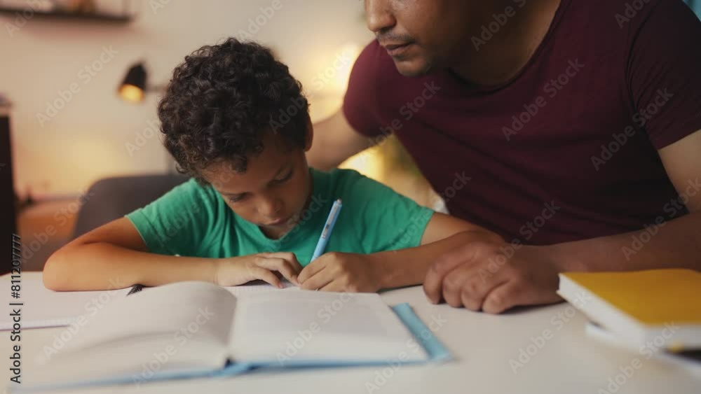Focused African American boy writes task in notebook, assisted by supportive dad Focused African American boy writes task in notebook, assisted by supportive dad