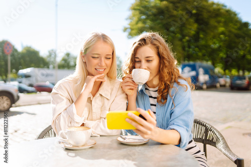 Two young women drinking coffee and using mobile phone while sitting at the summer cafe. Fashion, beauty, blogging, tourism.