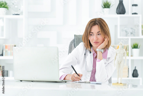 Portrait photo of young and beautiful female doctor in white medical gown sitting and working on laptop in the office of the modern clinic. Online consultation and distant cure concept © Vitaliy