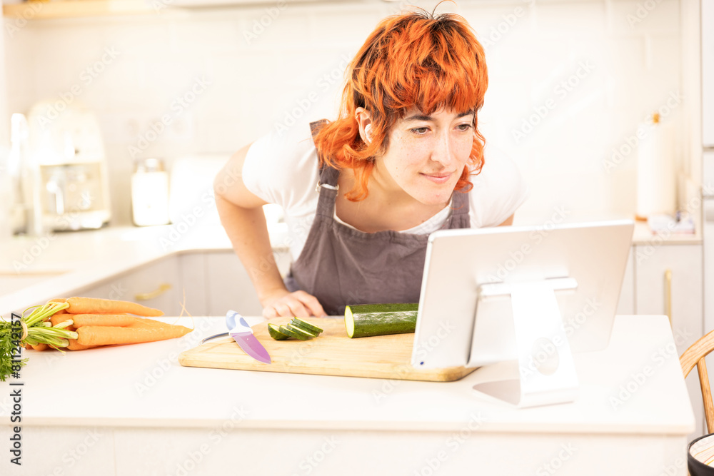 A woman is cutting vegetables on a cutting board while looking at a tablet. The tablet is displaying a recipe for a vegetable dish A woman is cutting vegetables on a cutting board while looking at a tablet. The tablet is displaying a recipe for a vegetable dish