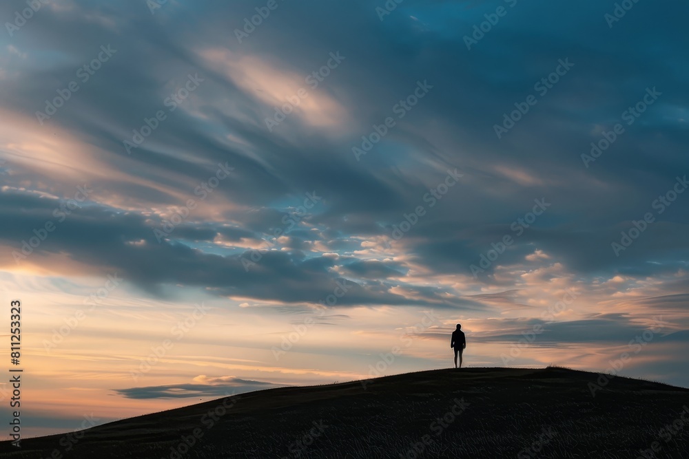 Fototapeta premium A person stands on a hilltop, looking out under a cloudy sky, A lone figure standing on a hilltop, watching the evening descend with a sense of peace and contemplation