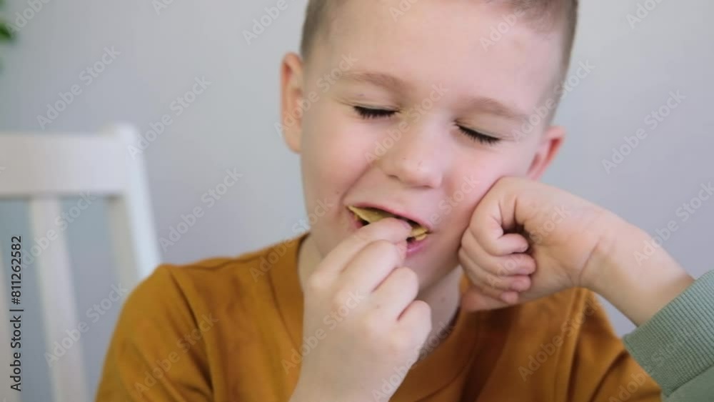Close-up of hungry child putting potato chips into his mouth while sitting at table. Boy happily eats fast food, junk food at home. Childhood obesity concept. Selective focus. Close-up of hungry child putting potato chips into his mouth while sitting at table. Boy happily eats fast food, junk food at home. Childhood obesity concept. Selective focus.