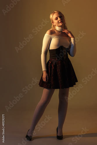 young blonde girl with bright makeup in black school dress poses as doll on yellow background in studio