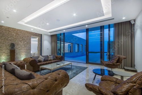 The luxurious interior of the living room with brown sofas and armchairs on the background of an open panoramic window. Evening Ocean View and Artistic Decor..