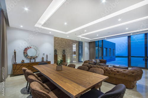 Modern Living Room With an expansive wooden dining table surrounded by plush leather chairs and Pool View During Evening. Large glass sliding doors offer a serene view of a swimming pool.