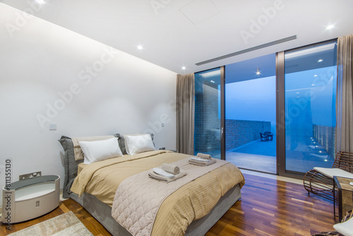 A luxurious bedroom with a king-size bed, white furnishings, panoramic window offering a beautiful view of the sunset.