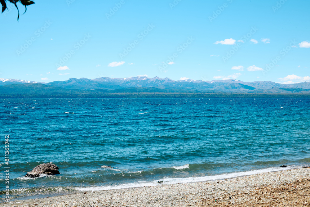 Obraz premium rocky shore of a blue lake, on the horizon a landscape of mountains with peaks covered with snow