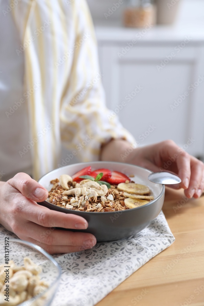 Woman eating tasty granola with banana, cashew and strawberries at wooden table indoors, closeup Woman eating tasty granola with banana, cashew and strawberries at wooden table indoors, closeup