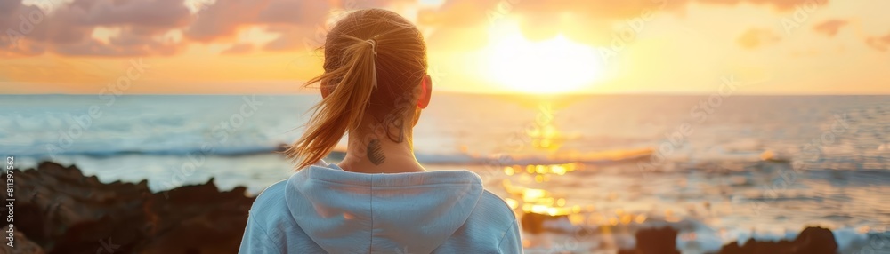 A sporty woman in a sweatshirt gazes out at the ocean, embracing the warmth of the setting sun A sporty woman in a sweatshirt gazes out at the ocean, embracing the warmth of the setting sun