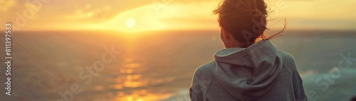 A sporty woman in a sweatshirt gazes out at the ocean, embracing the warmth of the setting sun photo