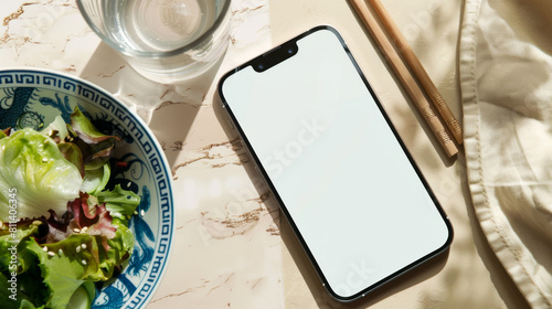 White Screen Smartphone Mockup with Salad and Chopsticks on Marble Table: Top View of Blank Screen Mobile Phone, Fresh and Healthy Salad and a Glass of Water