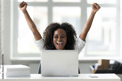 An overjoyed African American woman raising her arms in victory while sitting at a desk with a laptop, having won an online contest or gotten good news.