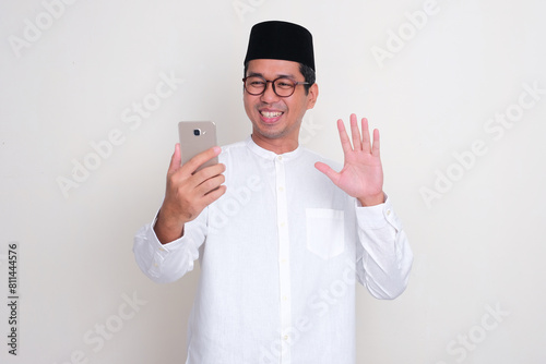 Moslem Asian man smiling and waving hand while video calling using mobile phone photo