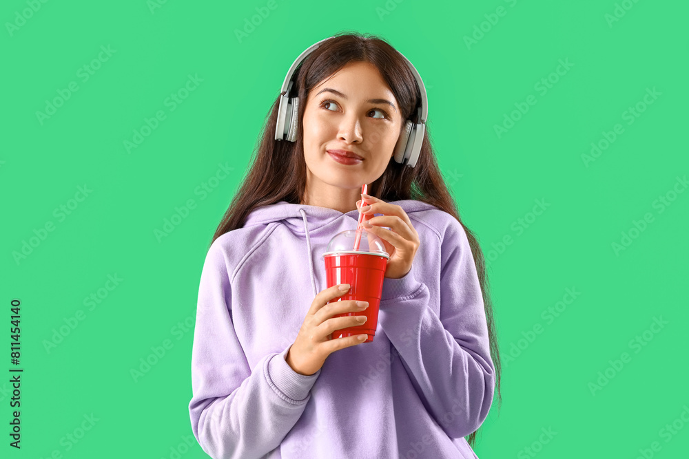 Beautiful young Asian woman in stylish hoodie and headphones holding cup of drink on green background Beautiful young Asian woman in stylish hoodie and headphones holding cup of drink on green background