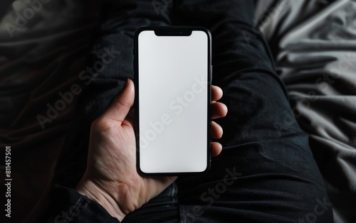 Mockup of a blank iPhone screen sitting on the lap, top view of a hand holding a phone with a white screen, wearing black pants and shirt with a dark background.