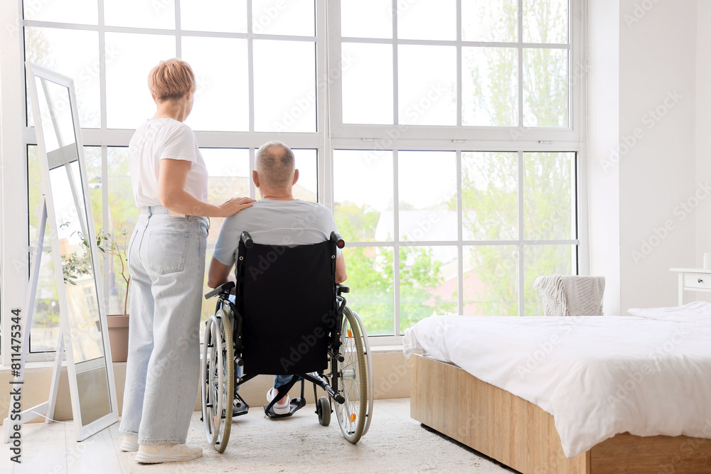 Mature woman with her sick husband in wheelchair at home, back view Mature woman with her sick husband in wheelchair at home, back view