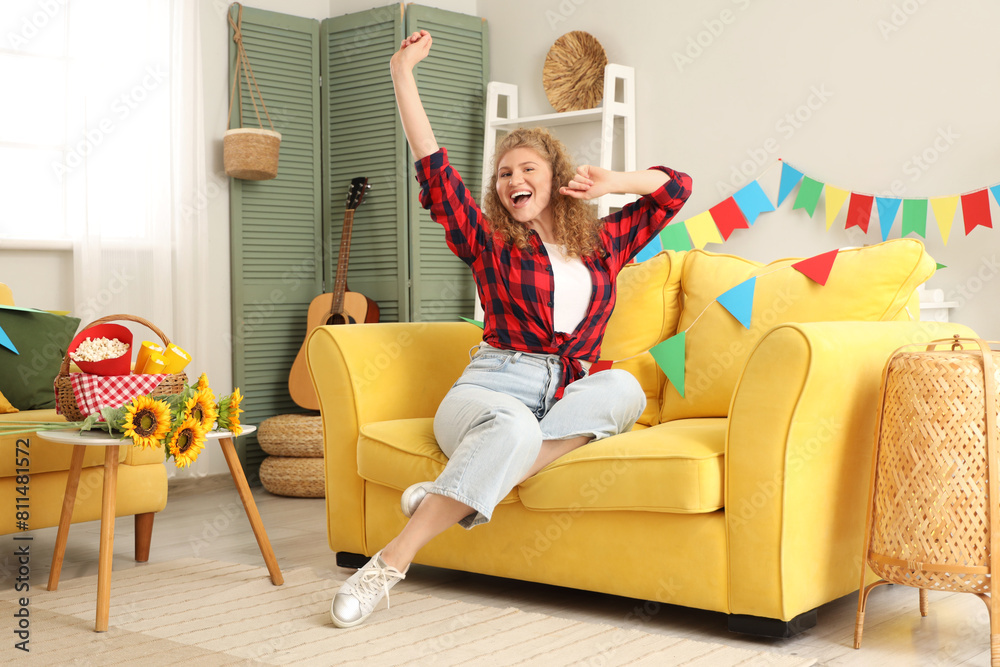 Happy young woman sitting on sofa with colorful flags at home. Festa Junina celebration