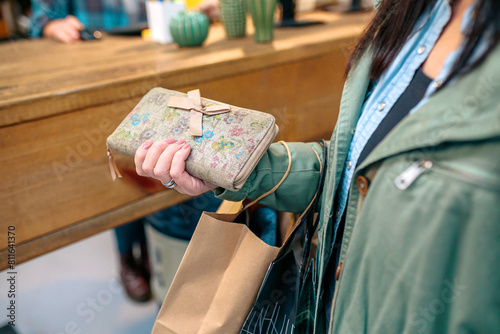 Close up of unrecognizable woman client holding wallet with bow and ecological shopping paper bags in front of a counter on local store. Female customer ready to pay purchased items on small business.