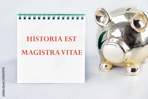 Historia est vitae magistra (History is the tutor of life) Latin phrase on a vertically standing notebook near the piggy bank on a light background photo