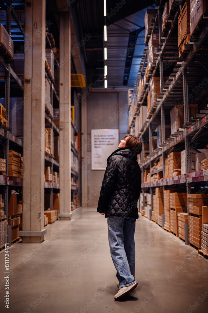 Young woman in a black puffer jacket and jeans, pausing to look up while walking through a warehouse. The vertical lines of the shelves 