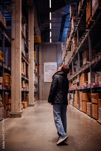 Young woman in a black puffer jacket and jeans, pausing to look up while walking through a warehouse. The vertical lines of the shelves and the overhead lighting draw the viewer's eye upwards