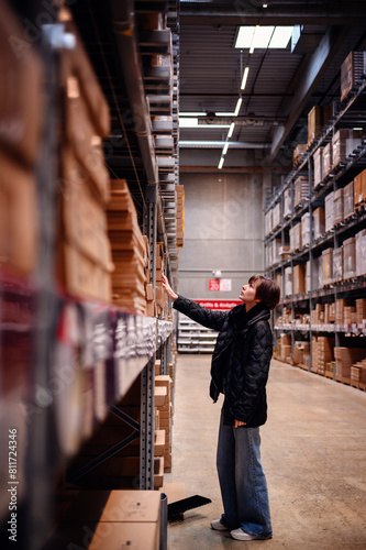 Young woman reaching towards a high shelf in a warehouse, illustrating her focus and determination. Her casual attire 
