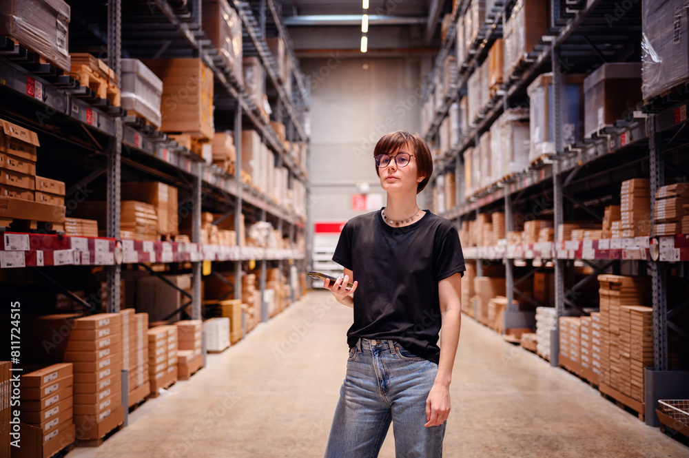 Young woman confidently walking down a warehouse aisle, smartphone in hand, possibly checking product locations or inventory details. Her modern, casual attire 