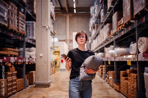 Young woman using her smartphone while shopping in a warehouse, holding a cushion in one arm. Her focused look at the phone screen 