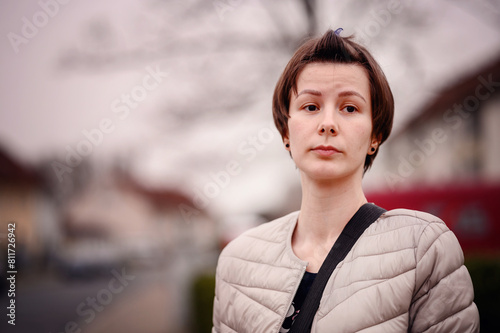A young woman with short brown hair gazes contemplatively while standing in a suburban neighborhood. She wears a light beige quilted jacket and carries a black shoulder strap,  photo