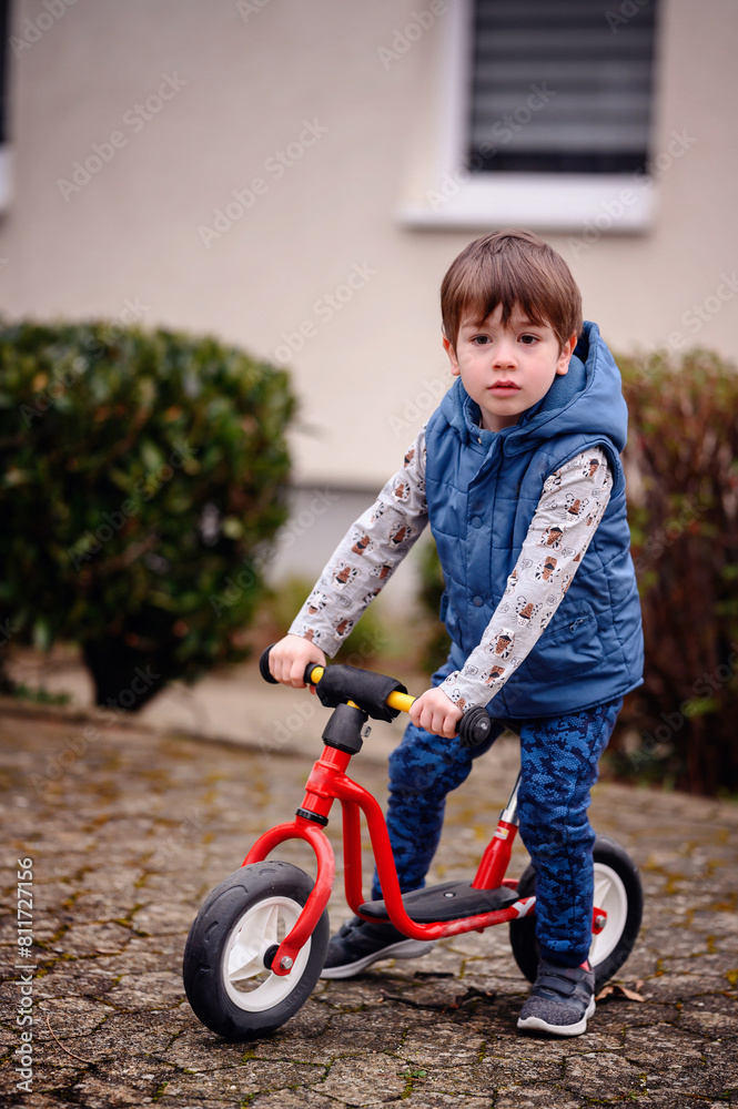 A focused young boy stands confidently with his red scooter, ready to embark on a ride. Dressed in a blue vest and a playful puppy-themed shirt, A focused young boy stands confidently with his red scooter, ready to embark on a ride. Dressed in a blue vest and a playful puppy-themed shirt,