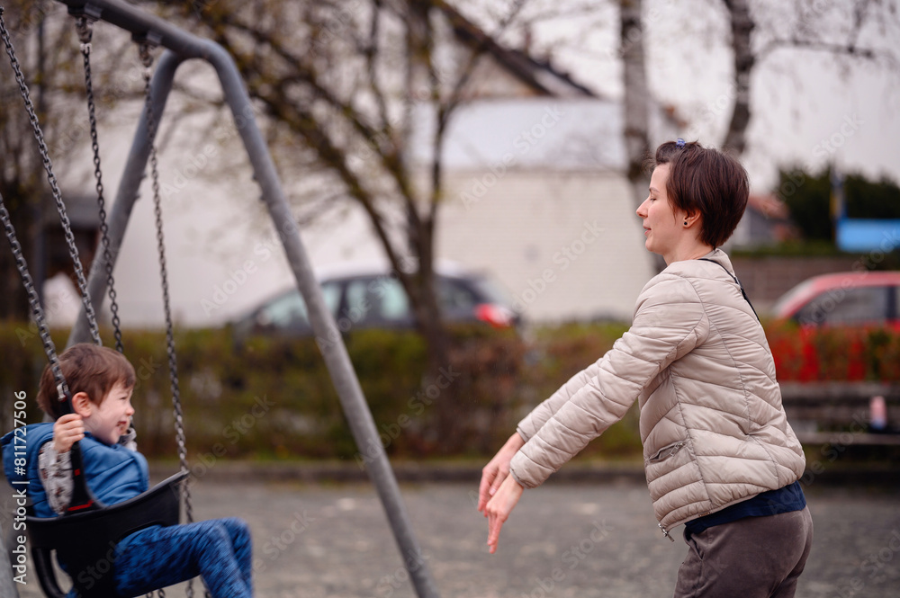 Obraz premium A heartwarming moment of a mother pushing her young son on a swing at a local park. The boy, laughing with glee, reaches out his feet to meet his mother's hands in a playful game. 