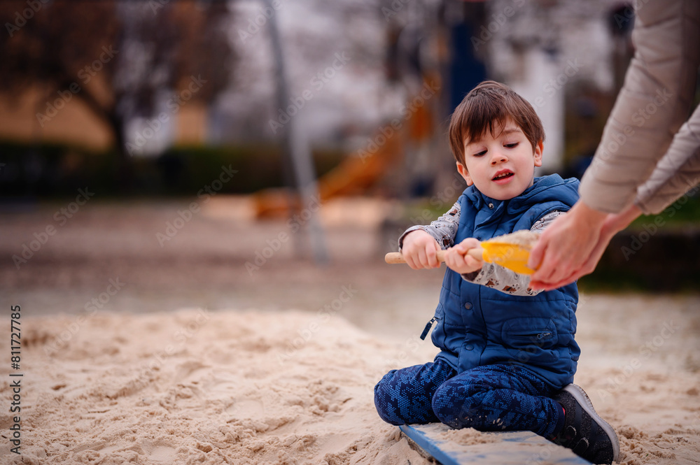 A young boy sits in a sandbox, deeply engaged in play. He holds a sandbox toy, lost in his creative world. His focused expression and the soft backdrop of the playground emphasize a peaceful, A young boy sits in a sandbox, deeply engaged in play. He holds a sandbox toy, lost in his creative world. His focused expression and the soft backdrop of the playground emphasize a peaceful,