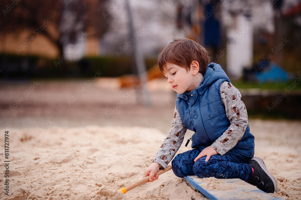 A young boy sits in a sandbox, deeply engaged in play. He holds a sandbox toy, lost in his creative world. His focused expression and the soft backdrop of the playground emphasize a peaceful A young boy sits in a sandbox, deeply engaged in play. He holds a sandbox toy, lost in his creative world. His focused expression and the soft backdrop of the playground emphasize a peaceful