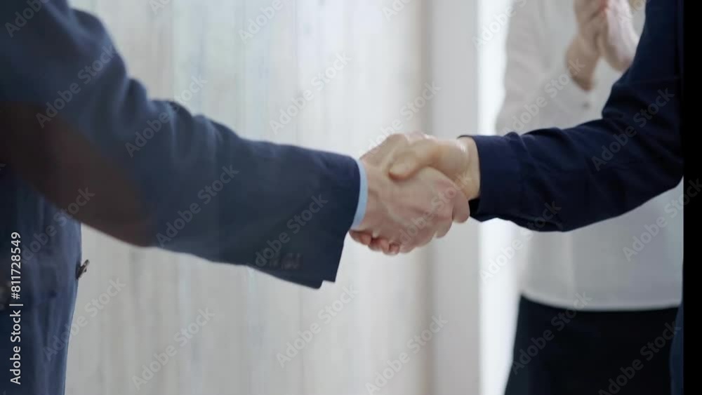 Close-up of a firm handshake between two professionals with a colleague applauding in the background. Close-up of a firm handshake between two professionals with a colleague applauding in the background.