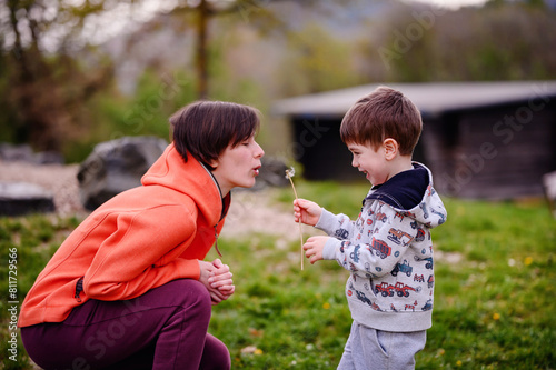 Φωτογραφία Captured in a serene natural setting, this image showcases a young boy holding a dandelion as he shares a moment with his attentive mother