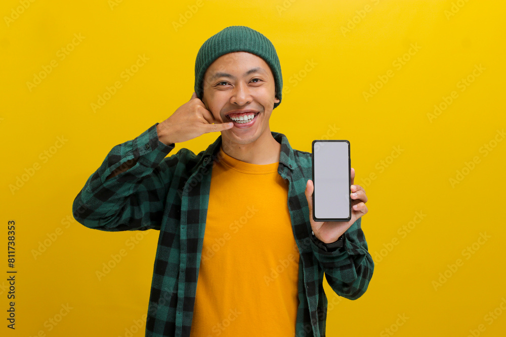 Cheerful Asian man in a beanie and casual shirt holds up his phone, screen facing the camera. He gestures a call me sign. Isolated on a yellow background. Communication, invitations, and contact. Cheerful Asian man in a beanie and casual shirt holds up his phone, screen facing the camera. He gestures a call me sign. Isolated on a yellow background. Communication, invitations, and contact.