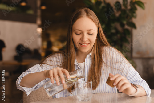 Woman pours water into a glass from a glass bottle. Blonde woman have a good time in cafe, she look happy and smiling. Girl wear stripped shirt.