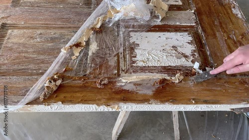 close-up of a caucasian man's hands scraping white paint off a wooden door with a metal scraper after it's been treated with chemical paint stripper photo