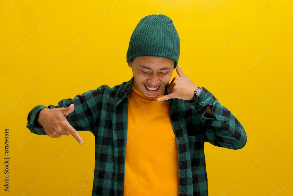 Enthusiastic young Asian man, dressed in a beanie hat and casual shirt, is gesturing a CALL ME while pointing down at an empty space, standing against a yellow background