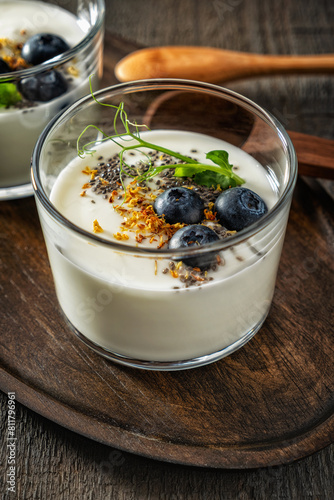 Homemade yogurt on a wooden tray with a wooden spoon on a textured gray wooden background, garnished with blueberries, chia seeds, dried osmanthus flowers and pea sprouts. Side and top view.