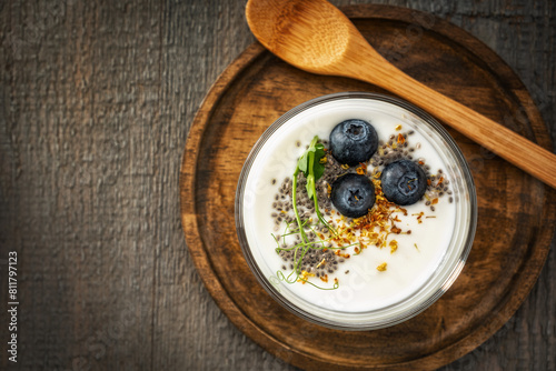 Homemade yogurt in a glass cup decorated with blueberries, chia seeds, osmanthus flowers and pea sprouts on a textured wooden plate with a wooden spoon, on a gray wooden background. View from above.